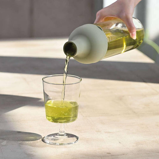 Hand die helder gele vloeistof uit groene fles schenkt in transparant glas op lichte tafel