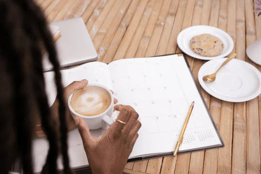 Persoon met koffie kopje voor agenda en gouden pen op houten bureau met koekje en laptop