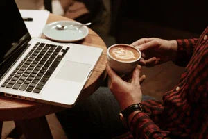 Persoon met geruit overhemd vasthoudt cappuccino bij laptop op houten tafel in kantooromgeving